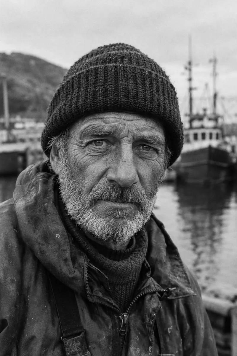 Black and white editorial portrait of a weathered fisherman with deep wrinkles and a knitted cap, looking straight into the lens, every pore and hair visible.